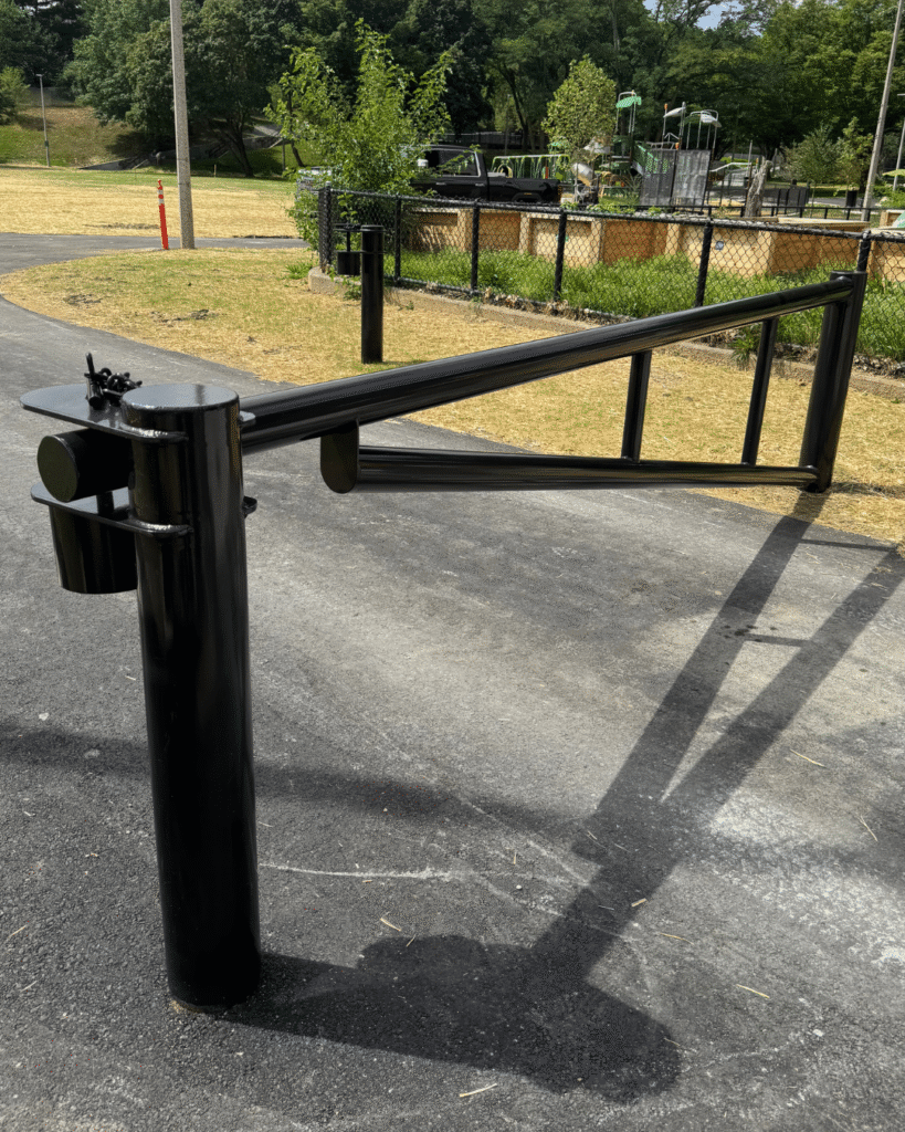 A black metal swing gate blocks a paved path in a park, with grass, trees, and fencing visible in the background on a sunny day.